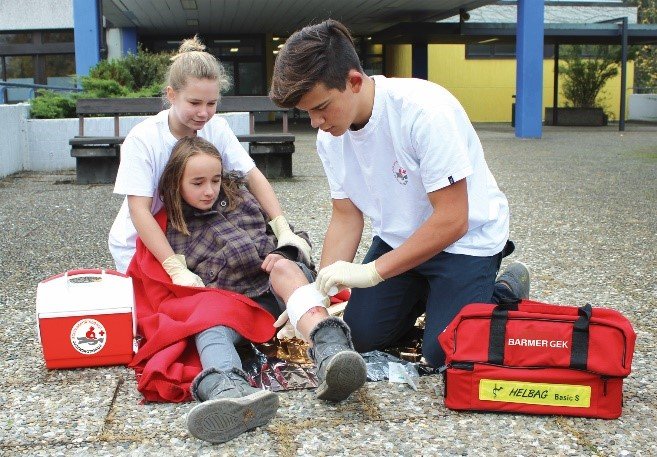 Eine Schulsanitäterin und ein Schulsanitäter helfen und beruhigen eine Mitschülerin, die mit einer Verletzung am linken Bein auf dem Boden liegt.