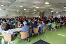Bild von der Begrüßung mit allen Teilnehmenden. Im Vordergrund sind anwesende Ehrengäste zu sehen (von hinten), im Hintergrund die Mensa der Schule, in welcher Schulsanitäts-Gruppen an Tischen sitzen.